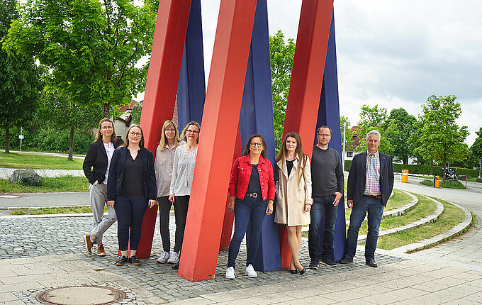 Gruppenfoto des Teams Forschung und Entwicklung vor der architektonischen Säule des B-Gebäudes im Sommer mit grünen Bäumen im Hintergrund 